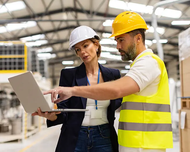 Responsabile tecnica e supervisore di fabbrica con elmetti protettivi che analizzano dati su un laptop in stabilimento industriale